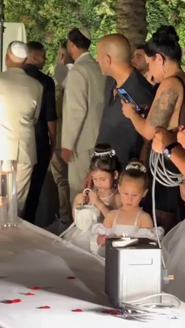 This video captures a gathering, likely a wedding, set outdoors at night. The scene is dimly lit, with a focus on a group of people and two young girls dressed in white flower girl dresses. The girls are adorned with pearl necklaces and hair accessories, suggesting a formal occasion. One girl is holding a small white bouquet, while the other appears to be looking at something in front of her.

In the background, several men are visible, some wearing yarmulkes, indicating a Jewish ceremony. The adults are dressed in formal attire, with one woman holding a smartphone. A metallic object, possibly a speaker or a piece of equipment, sits on a white-clothed table in the foreground, with red rose petals scattered around it. The overall atmosphere seems to be one of anticipation and celebration, with the soft lighting contributing to a somewhat intimate and festive mood. The presence of palm trees and dense greenery in the background suggests a warm climate, consistent with a location in Israel.
