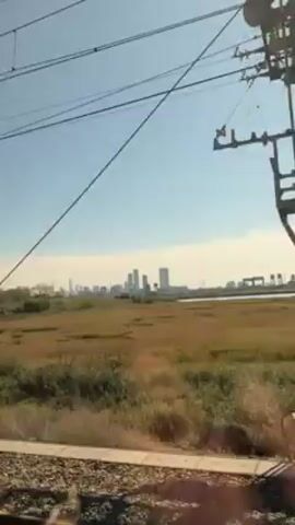 The video shows a view from a moving train. Outside the window, a landscape of a grassy marsh stretches out towards a skyline of buildings in the distance. Power lines are visible above.