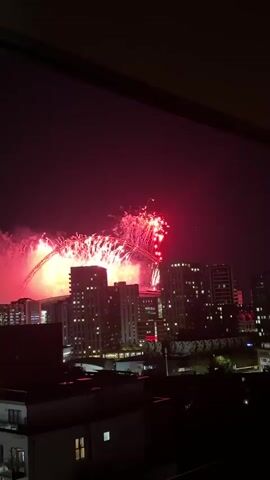 A nighttime aerial view of a cityscape during a fireworks display. The fireworks are primarily red and pink, illuminating the sky and reflecting off the buildings. The buildings are modern, with many windows lit up, suggesting a residential or office area. A large, arched structure, possibly a stadium or bridge, is visible in the background, partially obscured by the fireworks. The overall atmosphere is festive and celebratory.