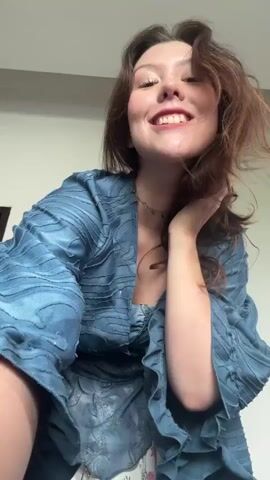 A young woman with long brown hair is dancing in front of a plain wall. She is wearing a blue, flowy top with wide sleeves and a layered skirt with floral patterns. Her arms are moving around her head and she is smiling. The lighting is soft and appears to be coming from above.
