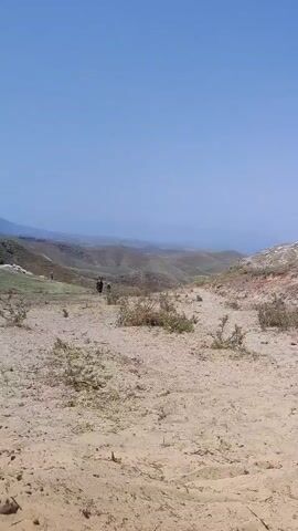 The video pans across a dry, sandy landscape with sparse vegetation. In the distance, a large, barren hill rises steeply. A few people are visible walking. There is a vehicle and a makeshift shelter in the mid-ground. A dog is seen moving in the foreground. There are also a few animals that appear to be donkeys or horses grazing on the hillside. The sky is clear and blue.