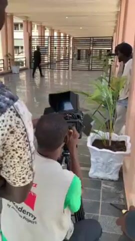 The scene depicts a film crew setting up a shot outdoors. A man dressed in black walks in the distance toward a gate. The primary focus is on a cameraman who is operating a professional video camera mounted on a tripod. He is wearing a white vest with "Film Academy by Jerusalem Project" printed on it. Another crew member stands next to him. A potted plant is positioned near the camera. The setting appears to be a tiled veranda, potentially part of a university or government building, given the architecture visible in the background.