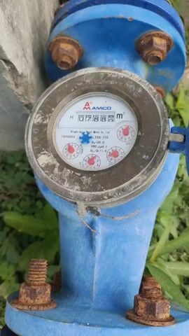 The video shows a close-up, eye-level shot of a blue water meter mounted on a pipe. The meter has a round, glass face with a digital readout and several analog dials. The digital display shows the number 0788. The analog dials, which have red needles, are also visible. The water meter is attached to a larger blue pipe with four rusty bolts. The background is out of focus but appears to be green foliage and a light-colored concrete surface. The lighting is natural and appears to be daylight. The video is approximately 5 seconds long and shows no discernible action beyond the static image of the water meter.