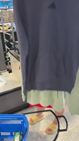 A young woman wearing orange-tinted sunglasses and a light-colored tank top is shown in a retail store. She is holding up two items of clothing on hangers. The first item, on the left, is a light green hooded sweatshirt with "Champion" written across the chest in a darker green color. The second item, on the right, is a red t-shirt with the Calvin Klein logo and text on it. The woman is holding both items up, presumably for inspection. In the foreground, a blue shopping basket is visible, containing what appears to be a green package. The background shows aisles of clothing racks, characteristic of a department store. The flooring is a speckled gray surface.