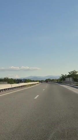 The video shows a first-person perspective of driving on a highway. The road is asphalt with a white line marking the lane. A white guardrail runs along the left side of the road. Trees and vegetation are visible beyond the guardrail, with hills or mountains in the distance under a clear blue sky with a few scattered clouds. Several cars are visible on the highway ahead.