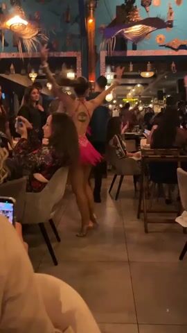 This video captures a lively evening scene inside a restaurant or club in Marrakesh, Morocco. The central figure is a woman performing a dance, dressed in a vibrant, two-piece hot pink costume with intricate embellishments and ruffles, paired with high-heeled shoes. She moves gracefully, twirling and raising her arms, captivating the audience.

Numerous patrons are seated at tables surrounding the dance area, engrossed in the performance. Many individuals are seen holding up their cell phones, actively recording the dancer, indicating their enjoyment and engagement. The setting is adorned with distinct Moroccan-inspired decor, including ornate hanging lamps, wall sconces, and decorative patterns in shades of blue and gold on the walls. The lighting is dim and atmospheric, suggesting it is nighttime. The overall ambiance is festive and entertaining, with people socializing and enjoying the cultural performance. No visible text is discernible in the scene.