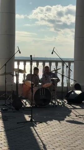 The video shows two people playing drums in front of a body of water. The sun is shining brightly, casting long shadows on the ground. There are microphones set up, suggesting a performance. One person is wearing a hat and appears to be moving around the drum set, while the other is seated and focused on playing. A bass guitar is visible to the left of the drum set. The scene is set outdoors, possibly on a promenade or pier, with large white columns framing the view. The sky is blue with scattered clouds.