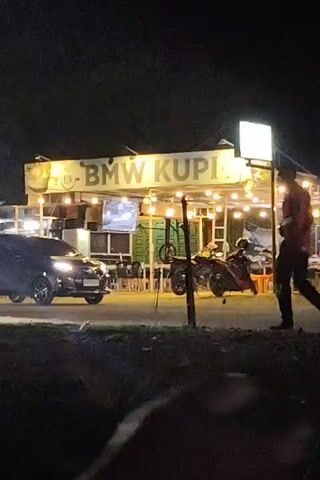 The video shows a nighttime scene with a building labeled "BMW KUPI" illuminated with lights. The building appears to be a cafe or restaurant with outdoor seating. A car drives past in the background, and a person walks across the road in front of the camera. Then, a scooter drives by on the road. The environment is dark, with some grass and foliage visible in the foreground. The focus is on the building and the traffic passing by.
