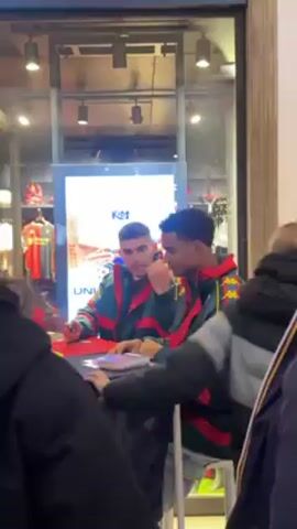 Two men are sitting at a table in front of a store window signing autographs. The men are both wearing matching green and red jackets with yellow trim. They appear to be interacting cordially, smiling at each other. People are lined up to the side of the table, waiting for autographs. The store window has a sign that says "KIDS" and displays images of clothing. The store is well-lit with recessed lighting.