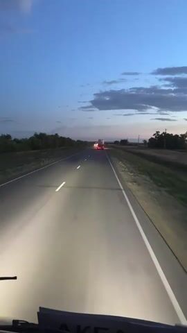 This media file captures a first-person perspective of driving on a highway at dusk, likely in Baiqadam, Kazakhstan. The scene is dominated by a two-lane asphalt road stretching into the distance, illuminated by the vehicle's headlights. The sky above is a deep blue, transitioning to lighter shades near the horizon, with scattered clouds suggesting a clear evening.

The primary subjects are the road itself and two trucks visible ahead. The truck in the foreground is a white semi-trailer, its brake lights glowing red. Further ahead, another truck, also a semi-trailer, is visible with its headlights on. The sides of the road are lined with sparse vegetation and trees, indicating a rural or semi-rural setting.

The activity taking place is vehicular travel. The time of day is clearly dusk or early evening, as evidenced by the blue sky and the use of headlights. The weather appears calm. There are no people directly visible, but the viewpoint implies a driver or passenger in the foreground vehicle.

Notable details include the intensity of the headlights, which create a bright pool of light on the road, and the blur of passing traffic, particularly a white van that overtakes the perspective vehicle. The overall mood is one of travel and solitude, with the vastness of the road and the fading light creating a sense of journey.

There is visible text on the dashboard of the vehicle in the foreground, partially obscured but appears to be the letters "AKE" in a dark font on a light background. There is also a small black camera mounted on the dashboard.