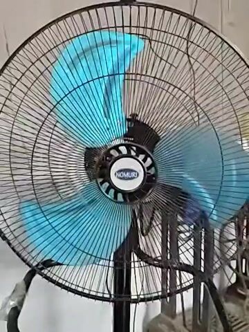 The video shows a Nomuri brand standing fan with blue blades rotating inside a black wire cage. The fan is on and positioned in front of a white or light-colored wall. Another fan is partially visible in the background.