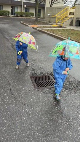 Two young children, dressed in matching blue rain suits and colorful rain boots, are seen walking on a wet, paved surface, likely an outdoor residential area or parking lot. The ground is visibly damp with scattered puddles, indicating recent or ongoing rain, and the sky appears overcast.

Each child holds a clear, dome-shaped umbrella featuring vibrant cartoon designs. The child on the left, walking towards the camera, has an umbrella with green, yellow, and red colors, and text like "ROAR" and "DINO" is visible, along with dinosaur figures. This child also holds a yellow object in their left hand, possibly a toy or part of the umbrella. The child on the right, initially walking away from the camera and then turning, has an umbrella with blue, green, and orange colors, also adorned with cartoon dinosaurs. Both children appear to be enjoying their time in the rain.

In the middle of the pavement, there is a dark, square metal storm drain cover. In the background, there are light-colored apartment buildings with multiple windows and balconies. To the right of the buildings, a bright yellow metal railing or staircase structure is visible. Some bare trees and bushes are also scattered in the background, suggesting a cooler season.