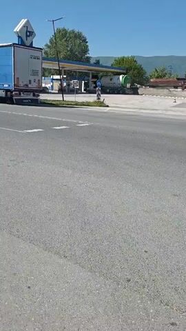 The camera is stationary and shows a view of a road and the surrounding area. Initially, a large blue and white semi-truck trailer is seen moving to the left. To the right of the trailer is a gas station. As the camera pans to the left, other trucks and cars can be seen on the road. A building with the word "CASINO" is also visible in the distance. There's also a restaurant or cafe with outdoor seating along the left side of the road. The overall impression is of a street scene in a commercial area during a sunny day.