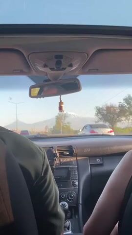 This video provides a first-person perspective from inside a car, looking forward through the windshield. The sun appears to be setting, casting a warm glow and long shadows, suggesting late afternoon or early evening. The weather seems clear, with a bright sky.

The primary subjects are the interior of the car and the landscape visible through the windshield. Inside the car, we see the dashboard with air vents, a rearview mirror hanging with a car air freshener, and parts of two passengers in the foreground – a person in a green jacket on the left and another person on the right whose arm is visible. The air freshener is a small glass bottle with red liquid, hanging from a wooden top.

Outside, the scene is dominated by a majestic snow-capped mountain in the distance, likely Mount Ararat, which is a prominent landmark in Armenia. The mountain is bathed in the golden light of the setting sun. In the middle ground, there are trees, utility poles with power lines, and a few other vehicles on the road, indicating that the car is in motion, likely driving on a road leading towards or away from the mountain. There are also some buildings visible, suggesting a rural or semi-urban setting.

The mood appears relaxed and scenic, with the beautiful vista of the mountain as the focal point. The visible text includes "SRS" on the dashboard, which likely refers to the Supplemental Restraint System (airbag system) of the car, and some Arabic or Persian script on the wooden cap of the air freshener. The location context of Ayntap, Armenia, further confirms the likely presence of Mount Ararat.