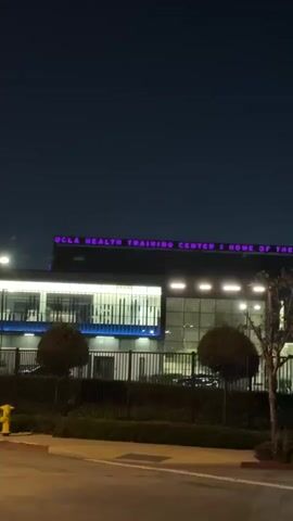 The video presents an exterior shot of a building at night. The sky is dark, indicating nighttime. In the foreground, there's a paved area, likely a road or sidewalk, with some small trees and bushes behind a fence. The main subject is a modern-looking building with several illuminated windows.

Prominently displayed on the top of the building in bright purple neon letters is the text: "UCLA HEALTH TRAINING CENTER | HOME OF THE LOS ANGELES LAKERS." This identifies the structure as the training facility for the Los Angeles Lakers basketball team, located in El Segundo, United States of America. The building itself is well-lit, contrasting with the dark night sky. No people or specific activities are visible, just the illuminated building and its surrounding landscape at night.