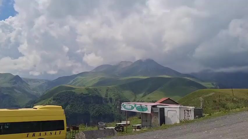 A yellow van with the word TRANSIT on its side is parked in front of a small roadside structure. The structure appears to be a food stand or shop, with a table and chairs set up outside. In the background, rolling green hills are visible, with dramatic, cloudy skies overhead. The mountains in the distance are partially obscured by clouds, adding to the atmospheric feel of the scene. The overall impression is one of a remote, scenic location.