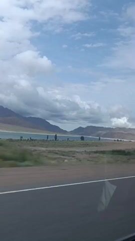 A road runs through a landscape with a body of water and mountains in the background. The sky is cloudy. The camera is moving.
