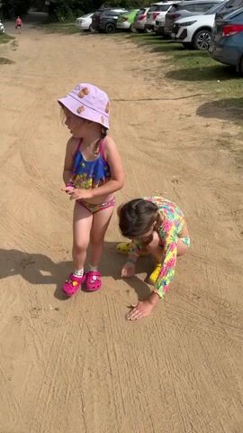 Two young girls are on a sandy path. The girl on the left wears a purple bucket hat and a blue and yellow swimsuit, with pink Crocs. She is standing and looking to her left. The girl on the right wears a brightly colored swimsuit and yellow Crocs. She is crouched down and appears to be looking at something on the ground. In the background, several cars are parked on grass. The scene is lit by daylight.