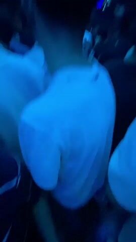 The video shows a crowded nightclub with a DJ booth and a large screen displaying a silhouette. People are dancing and enjoying themselves, illuminated by blue and white lights. The camera is positioned behind a person in a white t-shirt, facing the crowd. Several other individuals are visible, some with their backs to the camera. The atmosphere is energetic and lively, with a DJ presumably playing music.