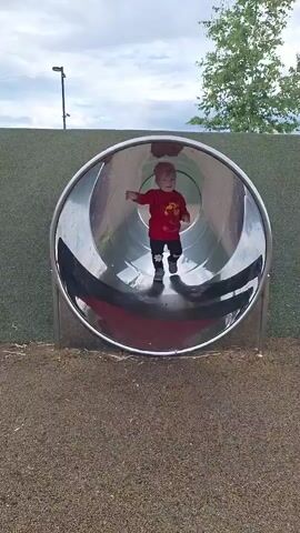 A young child wearing a red shirt, dark pants with floral designs, and a pink beanie emerges from a large, circular metal slide on a playground. The child is walking forward away from the slide. The playground surface is made of brown mulch. In the background, there is a green retaining wall, trees, and a cloudy sky with streetlights.