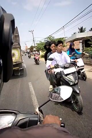 The video shows a first-person perspective from a motorcycle traveling on a road. The rider is wearing a black helmet and a dark jacket. In the foreground, the handlebar and a portion of the motorcycle's dashboard are visible. The road is paved and appears to be in a residential or commercial area, with buildings and trees lining the sides. Several other motorcycles and a truck are also seen on the road. One person is riding a red scooter, wearing a white helmet and a patterned shirt. Another person is riding a black scooter, wearing a dark hijab and a light blue backpack. A third person is walking on the side of the road, wearing a blue shirt. The sky is clear and the sun is shining. The video captures a dynamic street scene with multiple vehicles and pedestrians.