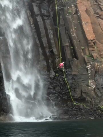 A person in a red jacket and white helmet rappels down a rocky cliff face next to a waterfall. A yellow rope is attached to the person and the cliff. The waterfall cascades down the rocks into a body of dark water below. The rock face is dark and has vertical striations.