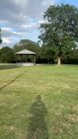 The video shows a park with a green lawn, surrounded by mature trees. In the middle distance, there is a gazebo with a light-colored roof. Two people are walking on the path behind the gazebo. The sky is partly cloudy with patches of blue. The shadow of the videographer is visible in the foreground of the shot. The video appears to be shot in daylight.