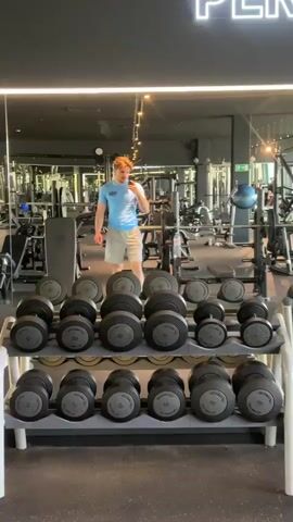 A young man stands in front of a dumbbell rack at a gym, taking a photo or video of himself with his cell phone. He is wearing a light blue t-shirt and light gray shorts. He is standing in a gym. There are multiple rows of dumbbells on the rack. Behind him, there are numerous weightlifting machines, benches, and exercise equipment scattered throughout the gym floor. The gym has mirrored walls, providing a sense of spaciousness. The lighting is bright, and the overall atmosphere appears clean and well-maintained.