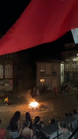 The video shows a nighttime scene in a cobblestone square. A large red flag or fabric is draped across the top of the frame, obscuring some of the view. In the center of the square, a bonfire burns, casting a warm glow on the surrounding area. Several people are gathered around the fire, sitting and standing. In the background, there are buildings with windows lit from within. Other individuals are seated in the background, some appearing to be watching the bonfire. The overall atmosphere seems to be one of a gathering or event taking place outdoors at night.