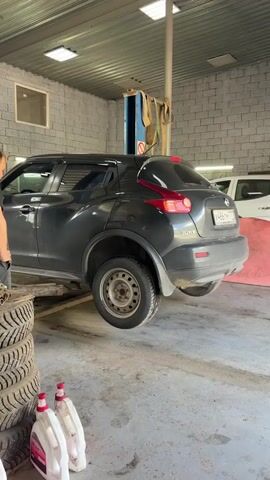 In a brightly lit garage, a dark gray Nissan Juke is elevated on a car lift, its rear passenger-side wheel suspended in the air. The car appears to be undergoing maintenance or repair. To the left of the vehicle, a stack of tires is visible, and two white and red plastic containers are on the floor. A person, partially visible from the side, stands near the front of the car, facing it. The garage walls are constructed of gray brick, and fluorescent lights are mounted on the ceiling. A blue and gray car lift is positioned behind the Nissan Juke.