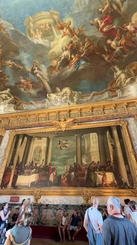 The video pans across a grand interior space with an elaborately decorated ceiling and a large painting on the wall. The ceiling is adorned with a fresco depicting numerous figures, likely angels and religious or mythological characters, set against a sky with clouds. The painting on the wall, framed in ornate gold, shows a scene with many people seated at long tables, suggesting a banquet or a significant gathering. Architectural elements like columns and arches are visible within the painting. In the foreground, a group of tourists are standing, looking up at the artwork. They are casually dressed and appear to be observing the art in a museum or historical building. Some are holding cameras or phones. The environment suggests an indoor public space, likely a museum, palace, or gallery, characterized by opulent decor.