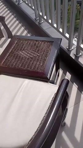 The video shows a balcony with a metal railing, a wicker table, and a view of a mountainous landscape. The sky is cloudy with the sun shining through. The camera pans up from the balcony furniture to the landscape and sky. The sun is visible through the clouds, creating a bright glare. The video ends with a view of the sun shining through the clouds.
