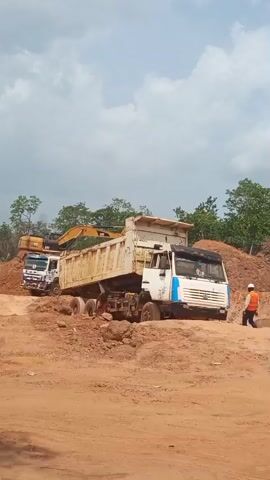 The video captures an active construction or excavation site in Aloko, Nigeria, under an overcast sky during the daytime. The ground is composed of reddish-brown earth, appearing dusty and uneven. Initially, a white dump truck, laden with a yellow excavator that has "CAT" visible on its arm, moves away towards the left of the frame. Another white dump truck is stationary in the foreground, with its dump bed slightly raised. Shortly after, an orange-yellow dump truck enters the scene from the right and drives across the open dirt area before coming to a stop. In the background, distant hills are visible beneath the cloudy sky, dotted with sparse trees and vegetation. The overall activity suggests earthmoving operations.