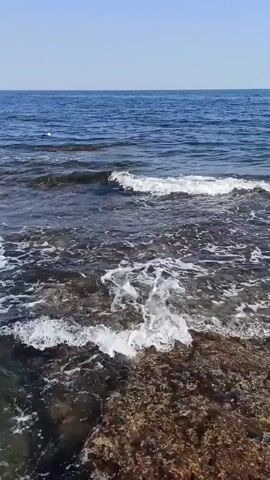 The video shows a rocky shoreline with waves crashing against the rocks. The water is clear and blue, and the sky is a light blue. The waves are breaking in the foreground, creating white foam as they hit the submerged rocks. In the background, the sea extends to the horizon under a clear sky. There is a small white object in the water in the distance, possibly a buoy or a small boat. The scene is peaceful and serene, capturing the natural beauty of the ocean.