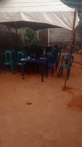 The video shows an outdoor gathering in a rustic setting. There is a makeshift tent providing cover over several plastic tables and chairs. Around the tables and chairs are several young children, some sitting and some standing. Behind them, women are working near a smoky cooking area, handling containers and preparing food. The ground is bare earth. The video is shot from a slightly low angle, panning across the scene.
