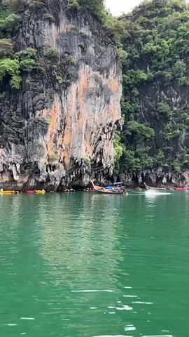 The video shows a body of green water with several longtail boats and kayaks. A large, rocky cliff face with lush green vegetation rises from the water on the right side of the frame. The cliff has streaks of orange and white rock. Several people are visible in and around the boats. One longtail boat is moving through the water, creating a wake. The water is calm with small ripples. The lighting suggests it is daytime, possibly overcast.