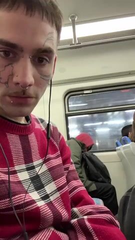 The video shows a close-up, first-person perspective of a person with face tattoos, a nose ring, and lip piercing. The person is wearing a red, black, and white plaid sweater and earphones. They appear to be on public transportation, likely a train, as indicated by the window view of a person sleeping and what looks like interior train seating. The lighting is dim, suggesting it is either nighttime or an overcast day. The person's gaze is directed towards the camera.