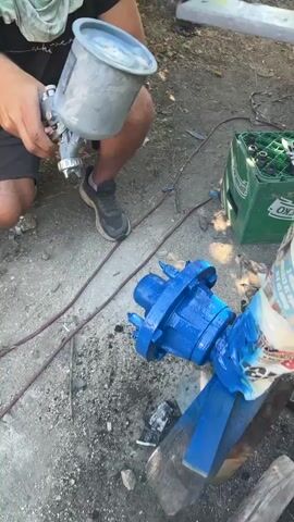 The video shows a person kneeling on the ground and painting a blue mechanical part with a spray gun. The person is wearing a black t-shirt, dark shorts, and grey athletic shoes. The mechanical part is mounted on wooden blocks. A green crate filled with bottles is visible in the background, along with various tools and debris on the ground. The environment appears to be outdoors.