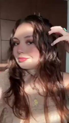 A young woman with light skin and brown, wavy hair looks at the camera in a selfie-style video. She wears a light brown top with lace detailing and a high neckline. She also has multiple gold necklaces, earrings, and a turquoise ring. The background is a plain, light-colored wall with some dark brown cabinets. The lighting is bright and sunny. She poses for the camera, running her fingers through her hair. She pulls a purse onto her shoulder.