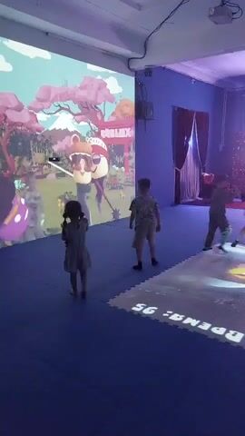 A group of children are playing a projected video game. The game appears to be a stylized cartoon world with trees, mountains, and buildings. The children are interacting with the game by moving their bodies, which are tracked by the projection system and translated into in-game actions. The room is dimly lit and the floor is covered in a blue mat. Some of the floor also has projected graphics on it, showing numbers and shapes. The children are dressed in casual clothing. One child is wearing a light-colored jumpsuit, another is wearing a patterned shirt and shorts, and a third child, who is mostly obscured, appears to be wearing a dark shirt and pants. The scene suggests an indoor entertainment or play area.