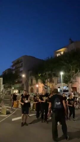 A group of people wearing black t-shirts with a green and red design are gathered on a street at dusk. They appear to be holding something in their hands, possibly instruments or percussion devices. One person in the foreground is facing away from the camera, wearing a black t-shirt and black pants, with a blue backpack. Another person in the foreground, facing the camera, is wearing a black t-shirt and black shorts, and has a beard. The street is paved, and there are buildings and trees in the background. The sky is a dark blue, indicating it is nighttime or late evening. Some of the people are looking at the person in the foreground facing away from the camera, who appears to be gesturing with their hand. The overall scene suggests an outdoor event or performance.