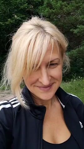 A woman with blonde hair and a black Adidas jacket smiles at the camera. The video is a close-up of her face and upper body. The background is blurry and appears to be outdoors, with trees and greenery visible.