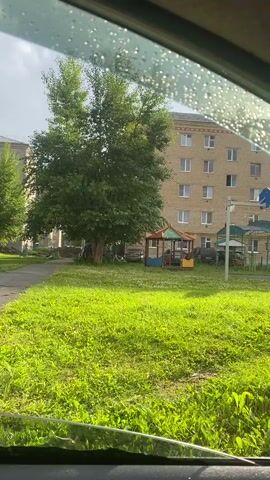This video, filmed from inside a vehicle looking out through a rain-streaked window, captures a residential scene in Uchaly, Russia. The primary subject is a multi-story, light-brown brick apartment building. In front of the building, a lush green lawn is prominent, with a large, leafy tree providing shade. A small, colorful playground structure, possibly a gazebo or playhouse, sits on the lawn near the tree. Other playground elements, like a blue basketball hoop mounted on a pole, are visible behind the smaller structure. Several cars are parked in the background, and a paved walkway runs along the edge of the grass. The sky appears overcast, suggesting a cloudy or rainy day, consistent with the water droplets on the car window. The sunlight, however, manages to break through, casting bright highlights on the grass. The overall atmosphere is one of everyday life in a residential area. No people are clearly discernible, and no text is visible in the frame. The time of day appears to be daytime, likely afternoon, given the angle of the light.