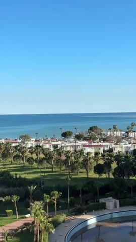 The scene presents a clear, sunny day with a view of the sea, a complex of white buildings, palm trees, and a drained circular pool in the foreground. The sky is a uniform, light blue. The sea extends horizontally, meeting the sky at the horizon. Along the coastline are closely packed white buildings interspersed with palm trees and some darker, rounded trees. The foreground showcases a grassy area dotted with more palm trees and shrubs, leading to a large, empty circular pool with a patterned bottom visible due to the lack of water. The overall impression is of a tropical resort area.