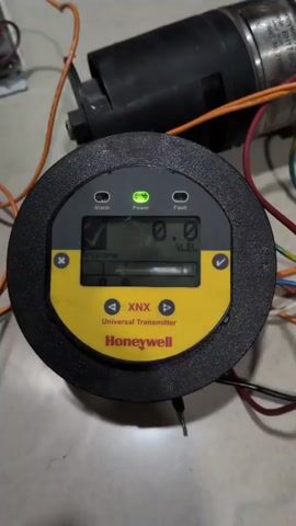 The video shows a close-up, overhead view of a Honeywell XNX Universal Transmitter. The device is circular with a black outer casing and a yellow and grey faceplate. The display screen shows "TOLUENE" on the left and "0.0 % LEL" on the right. There are indicator lights for Alarm, Power, and Fault, all of which appear to be off except for a checkmark next to Alarm. The device is connected to various wires and cables, suggesting it is part of a larger system. In the background, a black cylindrical object, possibly a motor or pump, is visible. The surface beneath the device appears to be tiled flooring. The lighting is somewhat dim, with a spotlight effect from above. There are no people visible in the video.