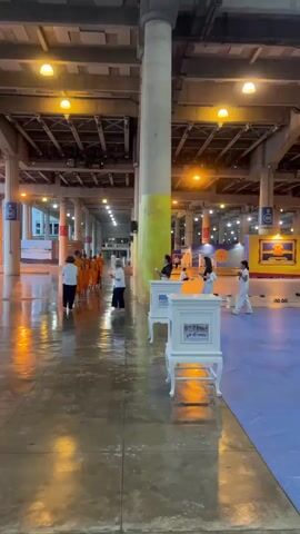 A group of monks in orange robes walk down a long hallway. The hallway has large concrete pillars and a high ceiling with lights. The floor is wet and reflects the lights. There are people standing and walking in the hallway, some are in white clothing. There are white pedestals with pictures on them in the hallway. The environment appears to be an indoor public space.