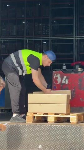 A male worker in a neon green safety vest, black t-shirt, black track pants, and a blue cap scans a white card using a barcode scanner gun. He stands on the loading dock, which is covered with a textured metal surface, in front of three cardboard boxes placed on a wooden pallet. To his left, another person is visible holding a white piece of paper. Behind them is a red forklift with the number "34" on it. The backdrop is a warehouse interior with multiple levels of dark shelves.