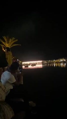 A night scene captures a body of water with a long, illuminated building across it. The building appears to be a restaurant or entertainment venue, lined with numerous lights that reflect on the water's surface. Several boats are moored in front of the building. On the left side of the frame, silhouetted figures are visible, standing near a palm tree. One person in the foreground is holding up a phone, seemingly recording the scene. The overall atmosphere is one of a lively evening, possibly a celebration or a popular tourist spot.