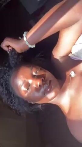 The video features a young, dark-skinned woman lying on her back. She has curly hair and is wearing a necklace and a bracelet. Her skin has a sheen, suggesting she may be wearing lotion or oil. She looks directly at the camera, occasionally sticking out her tongue. A smartphone is visible in the background. The lighting suggests the scene is bathed in sunlight.