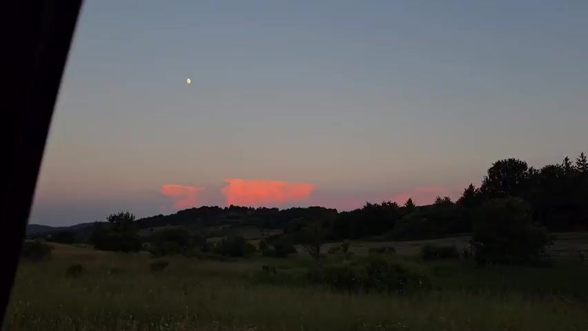 The video begins with a view of a rural landscape at dusk. The sky is a gradient of blue and pink, with a faint moon visible in the upper left corner. There are some brightly lit pink clouds over a line of trees in the distance. The foreground is a grassy field with scattered bushes. The camera is positioned as if looking out of a car window. The left side of the frame is dominated by the dark interior of the car.

At approximately 00:02, a person's face appears in the right side of the frame, looking out of the car window and smiling at the camera. The person's features are partially obscured by shadows and the angle. The video then returns to the exterior view of the landscape.